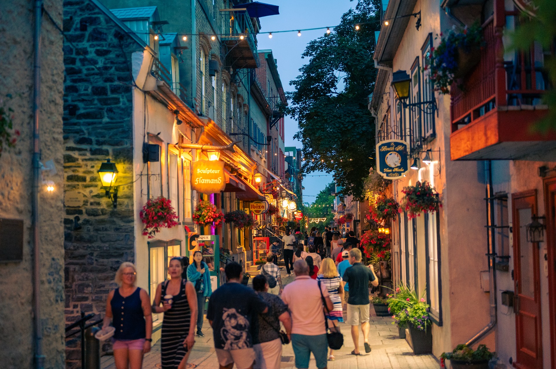 A lively narrow city street with warm shop lights at dusk